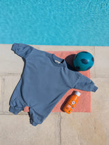 Sky-blue lightweight oversized baby bodysuit laid flat on a coral towel by a turquoise pool, next to a teal football and SPF 30 kids’ sunscreen.