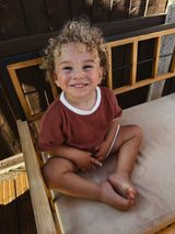 Smiling child with curly blonde hair wearing a cocoa brown French terry cotton T-shirt and shorts set with cream trim, sitting on a wooden bench.