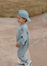 Child wearing a light blue outfit and cap standing on a concrete surface with grass in the background